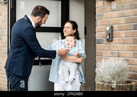 Casalinga con neonato che si trova di fronte alla porta d'ingresso della casa e saluta un marito e un papà dal lavoro. Foto Stock