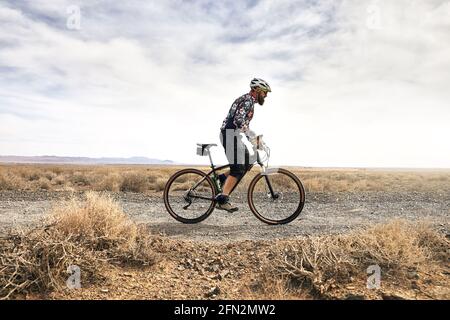 Il ciclista con la barba guida la sua mountain bike nella strada del deserto con il tempo soleggiato in Kazakistan. Sport estremi e attività ricreative all'aperto. Foto Stock