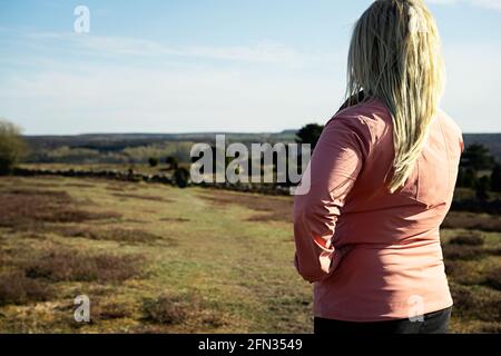 Donna con capelli biondi in una giacca a vento in piedi su un sentiero escursionistico in un campo, guardando il paesaggio paesaggio una giornata di sole a Brösarp, Svezia Foto Stock