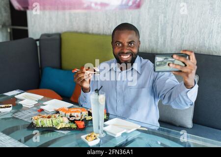 Eccitato sorridente giovane uomo d'affari africano che fa selfie shot o videochiamata sul telefono cellulare, tenendo chopsticks con sushi roll, con tradizionale pranzo orientale al ristorante Foto Stock