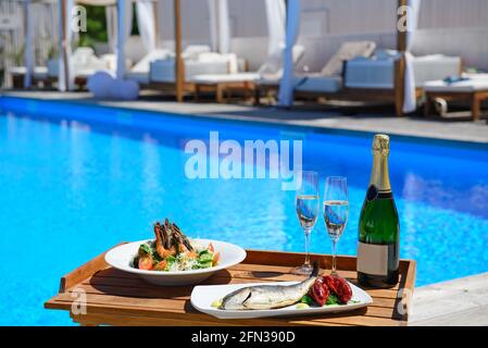 Champagne con due bicchieri di pesce e insalata di cesar servita a bordo piscina su vassoio di legno. Stile di vita di lusso. Foto Stock