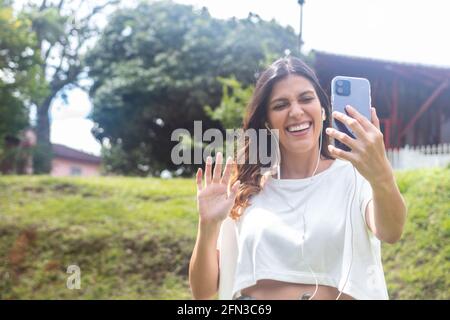 Donna latina con cuffie che prendono un selfie alzando la mano e sorridendo durante una videochiamata Foto Stock