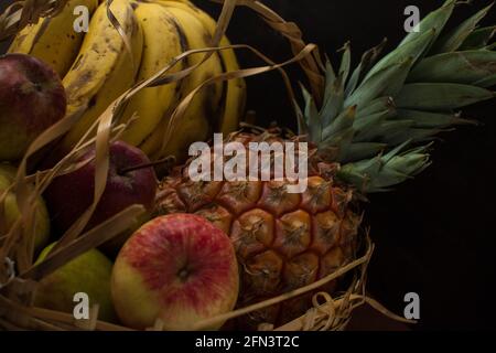 Alcune banane, mele e un ananas in un cesto su un tavolo nero con fondo nero Foto Stock