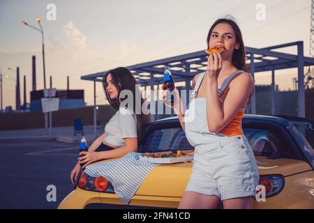 Belle Signore in abiti casual mangiare pizza, gustando acqua soda in bottiglie di vetro, in posa vicino auto gialla sul parcheggio. Primo piano, spazio di copia Foto Stock