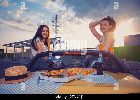 Le femmine degli amici sorridono, ponendosi in cabina gialla con patatine fritte, pizza e soda in bottiglie di vetro sul suo tronco. Fast food. Spazio di copia Foto Stock