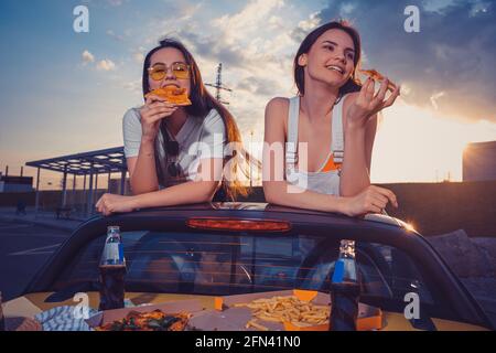 Le donne affascinanti stanno mangiando la pizza e sorridendo mentre posano in automobile gialla con le patatine fritte e la soda in bottiglie di vetro sul tronco. Fast food. Spazio di copia Foto Stock