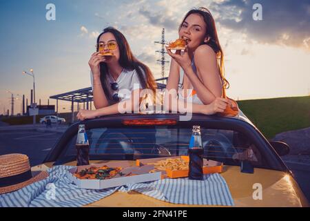 Le donne di charme stanno mangiando la pizza mentre si posa in cabina gialla con patatine fritte e soda in bottiglie di vetro sul tronco. Fast food. Primo piano Foto Stock