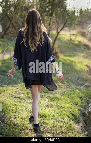 Bella ragazza bionda che cammina sull'erba. Una giovane donna con un abito elegante vaga in un parco circondato dalla natura durante un tramonto. luce calda e pace Foto Stock
