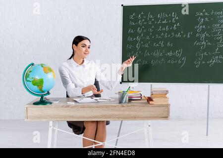 Insegnante sorridente che punta alla lavagna vicino al globo e libri dentro in aula Foto Stock