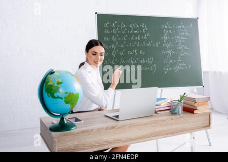 Insegnante sorridente che punta alla lavagna durante la lezione online sul laptop vicino globo e libri Foto Stock