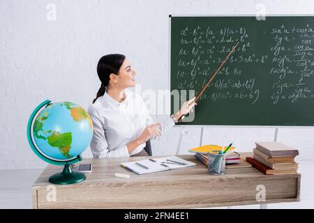 Insegnante sorridente che punta alla lavagna con le equazioni vicino ai libri e. globo a scuola Foto Stock