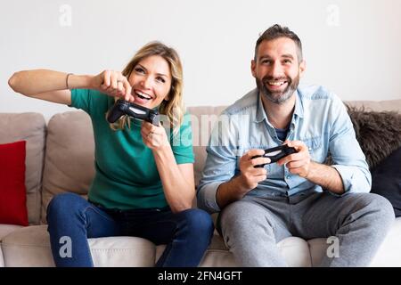 La coppia felice gioca alla console con i joystick. Gli amanti simpatici giocano con energia e divertimento. Le persone sorridenti giocano insieme durante il periodo di blocco. Foto Stock