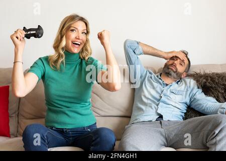 La coppia felice gioca alla console con i joystick. La donna bionda vince il gioco e si rallegra. Il marito triste e stanco crolla sul divano. Foto Stock