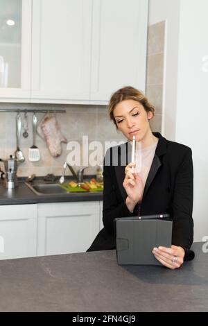 Attraente donna d'affari pensa di tenere un tablet. Una donna lavora da casa sul tavolo da cucina. Ragazza classy in vestito chiedendosi circa che cosa scrivere. Foto Stock