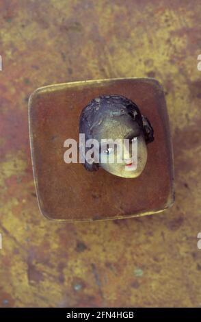 Testa della bambola degli anni '20 o ornamento con carnagione di olive e capelli neri giacenti su un piccolo contenitore di ottone Foto Stock