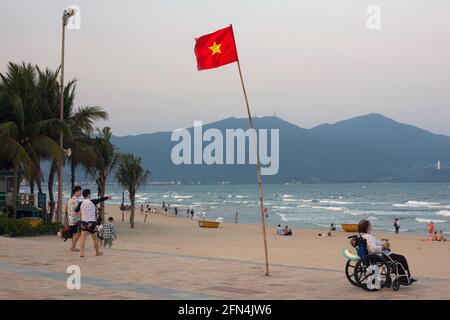 Bandiera del Vietnam che vola e persona in sedia a rotelle osservare le persone su My Khe Beach, da Nang, Vietnam Foto Stock