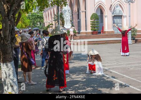 Attraente turista femminile che indossa ao dai e cappello conico fa segni di pace con le braccia distese alla Cattedrale di da Nang (chiesa rosa), da Nang, Vietnam Foto Stock