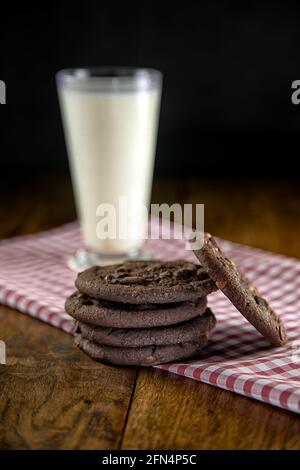 Pila di biscotti al cioccolato su panno rosso di gingham con vetro di latte su un vecchio tavolo di legno Foto Stock