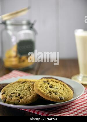 Pila di biscotti al cioccolato su panno rosso di gingham con vetro del latte e del vasetto di biscotti Foto Stock