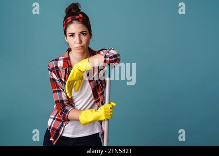 Annoiato attraente donna che indossa grembiule e guanti di gomma in piedi con un mop isolato su sfondo blu Foto Stock