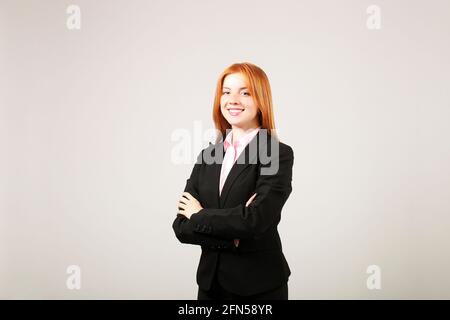 Giovane bella donna d'affari sicura che indossa vestito nero in piedi con le braccia incrociate sul petto e sorridente. Attraente femmina a testa rossa in noi formale Foto Stock