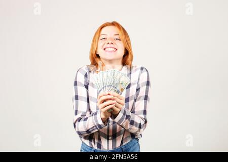 Ritratto di giovane bella donna rossa con pugnale di soldi che tengono uno stack di bolletta di cento dollari come ventilatore. Eccitata attraente rosso capelli femmina con/ Foto Stock