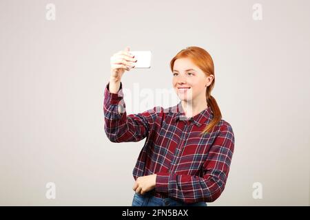 Ritratto di giocosa donna rossa con camicia a scacchi in flanella che prende un selfie con telefono cellulare, isolato sfondo grigio. Studentessa in possesso Foto Stock