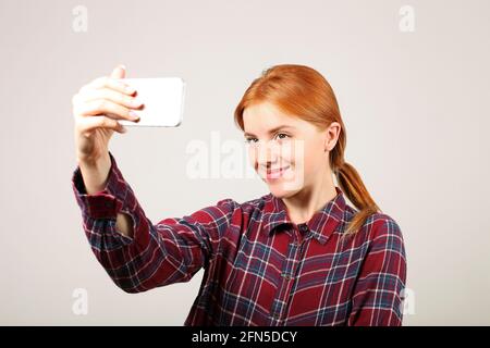 Ritratto di giocosa donna rossa con camicia a scacchi in flanella che prende un selfie con telefono cellulare, isolato sfondo grigio. Studentessa in possesso Foto Stock