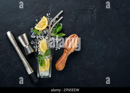 Mojito o cocktail limonata in calice con cubetti di ghiaccio, menta e lime su fondo di pietra nera con agitatore e bicchiere. Barra dei menu. Spazio per il testo Foto Stock