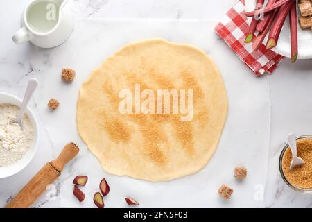 Home Baking concept con pasta cruda arrotolata con ingredienti farina, acqua, burro, zucchero e rabarbaro per la cottura su fondo di marmo bianco. Vista dall'alto Foto Stock
