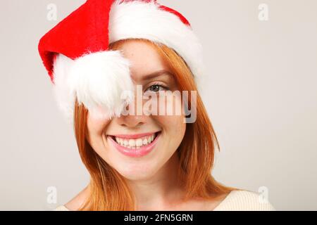 Primo piano ritratto di bella giovane donna rossa con cappello Babbo Natale e maglione bianco con espressione facciale soddisfatta. Carina femmina che festeggia Foto Stock