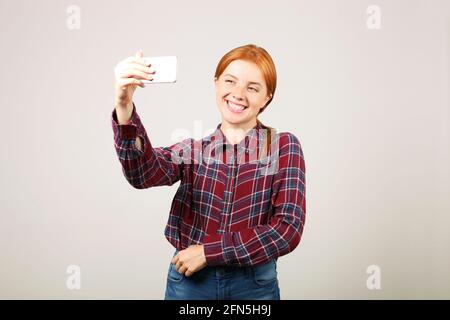 Ritratto di giocosa donna rossa con camicia a scacchi in flanella che prende un selfie con telefono cellulare, isolato sfondo grigio. Studentessa in possesso Foto Stock