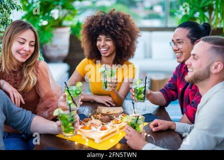 Gruppo multirazziale di amici che si godono mojito in un bar ristorante al tramonto in estate facendo un brindisi. I giovani tengono un cocktail con il cibo Foto Stock