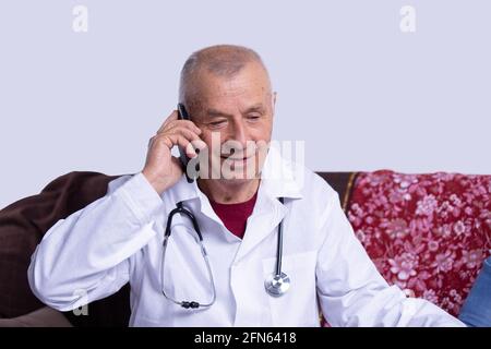 Ritratto di un medico anziano, un uomo dai capelli grigi che prende una pausa seduto sul divano, utilizzando il telefono cellulare. Vestendo abito bianco e stetoscopio. Coronavirus covid Foto Stock