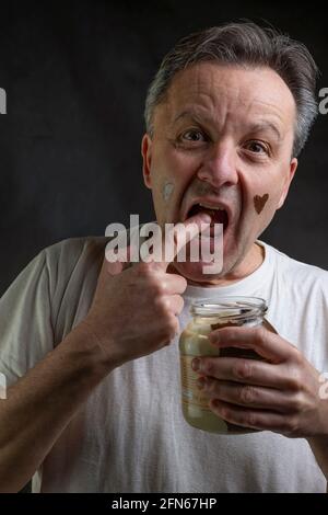 Un uomo di mezza età mangia avidamente una crema di cioccolato con la sua dito dritto dal vaso di vetro che rende un molto divertente espressione Foto Stock