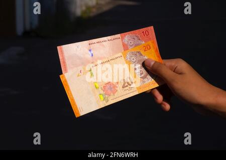 Mano asiatica del bambino che tiene 10 e 20 soldi della Malesia di Ringgit, fuoco basso, su sfondo scuro Foto Stock