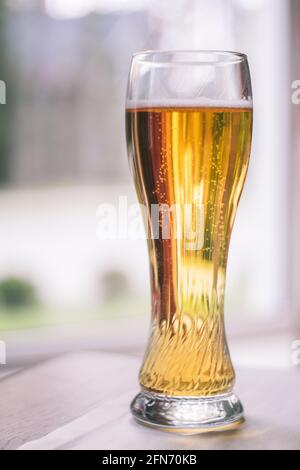 Bicchiere di birra chiara parzialmente sfocato con bolle, su tavolo di legno con tovaglia bianca vicino alla finestra. Verticale Foto Stock