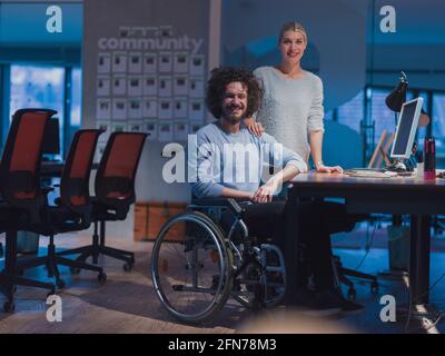 Uomo d'affari in sedia a rotelle in un moderno spazio ufficio di lavoro lavoro di notte in ufficio. Colleghi in background. Concetto di disabilità e handicap. Foto Stock