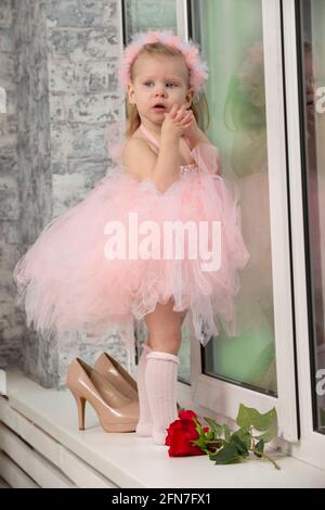 Bambina divertente in un elegante abito rosa e tacchi alti vicino alla finestra. Foto Stock