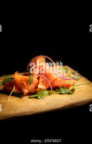 Salmone affumicato affettato e arrangiato con rucola un antipasto popolare, su un piatto di legno fine uno sfondo nero Foto Stock