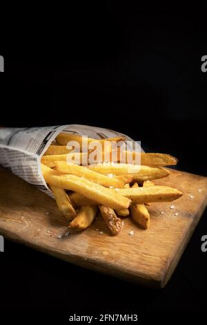 Patatine fritte con sale marino grossolano in un sacchetto servito su una tavola di legno e sfondo nero Foto Stock