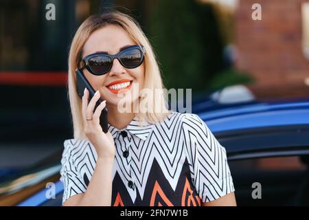 Donna che utilizza telefono cellulare, comunicazione o applicazione online, in piedi vicino all'auto sulla strada della città o parcheggio, all'aperto. Servizio di autonoleggio Foto Stock