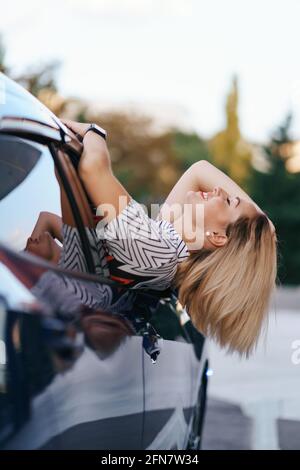 Allegra donna caucasica guida attraverso la pittoresca città soleggiata e. ondeggia le braccia mentre si allunga fuori dal finestrino dell'auto in una bella giornata Foto Stock