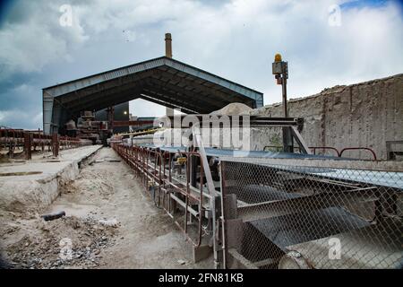 Temirtau, Kazakhstan: Vecchio cementificio sovietico. Nastro trasportatore in gomma sui rulli. Tetto in metallo e macchina di carico sullo sfondo. Cielo blu, nuvole. Foto Stock