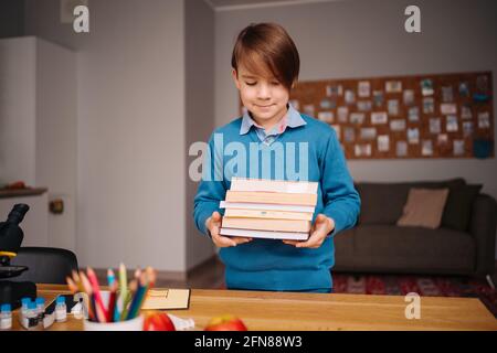 Formazione online a distanza, apprendimento su Internet. Ragazzo di primo grado che studia a casa, tiene un mazzo di libri, preparandosi per la lezione online Foto Stock