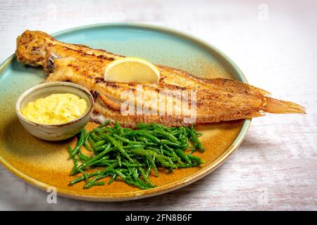 Pesce di passera intero al forno servito su un piatto colorato con limone, samphire e salsa di burro Foto Stock