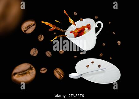 Tazza di porcellana bianca levitante con spruzzi di caffè da essa, cucchiaio, piattino circondato da chicchi di caffè su sfondo nero Foto Stock