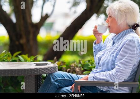 Attraente donna di mezza età alla moda che si rilassa con una tazza di caffè o tè ad un tavolo nel giardino in a. primo piano della vista ritagliata ad angolo basso Foto Stock