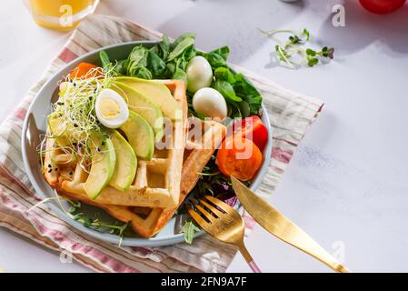 Waffle belgi appena sfornati con spinaci, uova, pomodori e avocado su piatto blu. Waffle salati. Colazione sana. Foto Stock