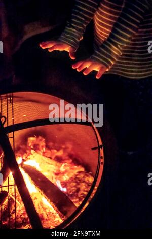 Il bambino riscalda le mani di fronte ad una fossa di fuoco Foto Stock